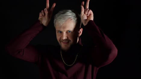 Funny-joyful-man-in-stylish-blouse-making-bunny-ears-hand-gesture-over-head-on-black-background