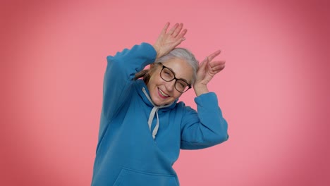 Funny-silly-senior-old-granny-woman-smiling-friendly-and-doing-bunny-ears-gesture-on-head,-fooling