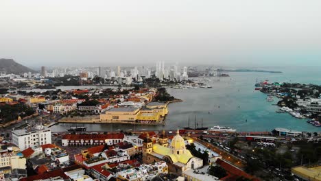 Vista-Aérea,-Seguimiento,-Toma-Aérea-Con-Drones-Hacia-La-Iglesia-Parroquial-San-Pedro-Claver-Y-El-Puerto,-Con-El-Centro-De-Cartagena-Al-Fondo,-Durante-La-Puesta-De-Sol,-En-Colombia,-América-Del-Sur