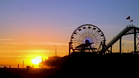 Hermoso-Atardecer-Naranja-Con-Lapso-De-Tiempo-De-La-Noria-Y-Paseos-En-La-Parte-Superior-Del-Famoso-Muelle-De-Santa-Mónica-En-La-Playa-De-California-Con-Banderas-Ondeando-Al-Viento-Y-Gaviotas-Volando-Alrededor