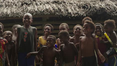 Establishing-close-shot,-indigenous-kids-jumping-and-smiling-around,-wooden-Hut-in-the-background