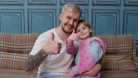 Father-and-little-child-daughter-kid-in-pajamas-sit-on-couch-in-room-smiling,-showing-thumbs-up