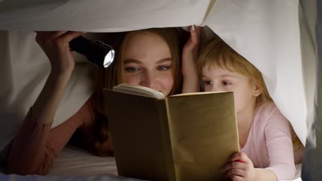 Mother-and-daughter-lying-in-bed-at-home-under-duvet-blanket-and-reading-bedtime-stories,-fairytales