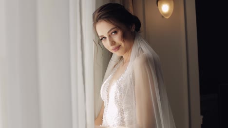 Smiling-bride-in-white-dress-staying-near-window,-looking-at-camera-wedding-preparations-at-hotel