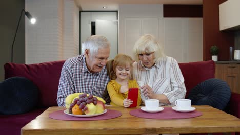 Grandparents-teaching-small-kid-granddaughter-using-smartphone-making-online-shopping-at-home