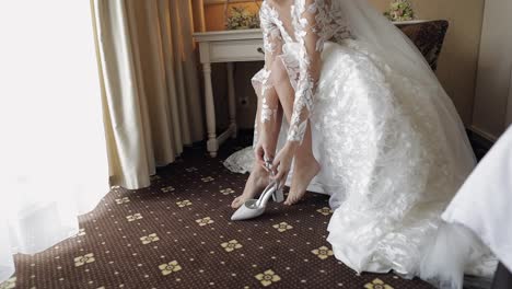 Young-bride-in-white-wedding-dress-wearing-high-heeled-shoes-on-slim-legs-while-sitting-at-home