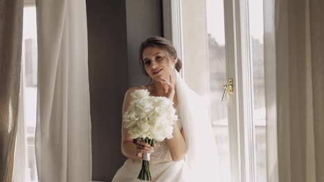 Bride-in-white-dress-staying-near-window-with-flowers-bouquet,-wedding-morning-preparations-at-home