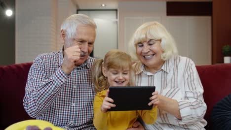 Senior-Caucasian-couple-with-cute-child-girl-granddaughter-using-tablet,-watching-funny-videos