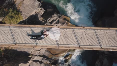 Frischvermählte.-Braut-Und-Bräutigam-Liegen-Auf-Einer-Brücke-über-Einem-Gebirgsfluss.-Luftaufnahme