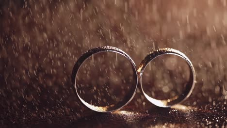 Two-gold-wedding-rings-under-splashes-of-water-on-dark-surface-shining-with-light-close-up-macro