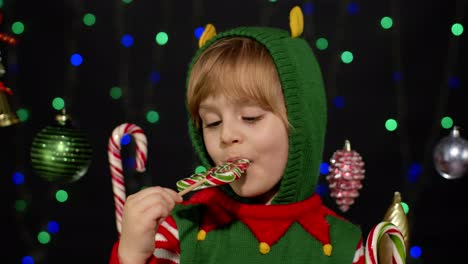 Child-kid-girl-in-Christmas-elf-Santa-Claus-helper-costume-licking-candy-lollipop-caramel-sweets