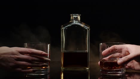 Two-hands-with-glasses-of-cognac-whiskey-with-ice-cubes-making-cheers-toast-on-black-background