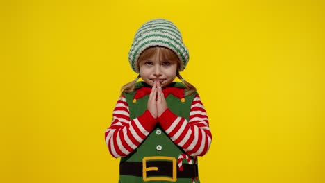 Kid-in-Christmas-elf-Santa-helper-costume-begs,-prays-about-gifts-on-New-Year.-Keeps-palms-together