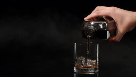 Barman-pour-whiskey-cognac-brandy-from-bottle-into-drinking-glass-with-ice-cubes-on-dark-background