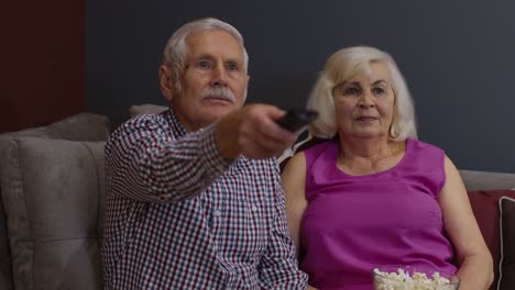 Senior-old-couple-holding-remote-control-talking-laughing-watching-humor-tv-show-sitting-on-sofa