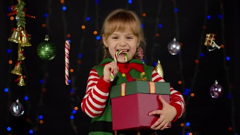 Kid-girl-in-Christmas-elf-Santa-Claus-helper-costume-with-gift-box-licking-candy-cane-lollipop