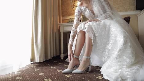 Young-bride-in-white-wedding-dress-wearing-high-heeled-shoes-on-slim-legs-while-sitting-at-home