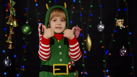 Teen-kid-girl-in-Christmas-elf-Santa-Claus-helper-costume-showing-thumbs-up-on-black-background