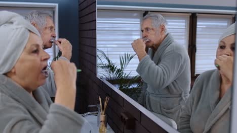 Senior-couple-grandmother-and-grandfather-brushing-teeth-and-looking-into-a-mirror-at-bathroom