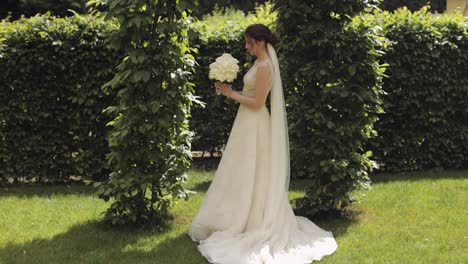 Beautiful-stylish-bride-in-white-wedding-dress-and-veil-holding-wedding-bouquet-in-hands-in-park