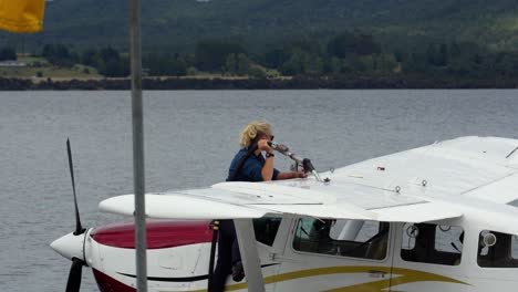 Female-pilot-stands-on-seaplane-at-water-harbor-port-refueling-plane-at-water's-edge,-plane-bobbing-in-the-water