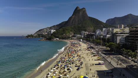 AERIAL:-crowded-Praia-do-Leblon-beach,-sunny-day-in-Rio-de-Janeiro,-Brazil