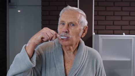 Senior-man-grandfather-in-bathrobe-brushing-teeth,-looking-into-mirror.-Morning-hygiene-at-bathroom