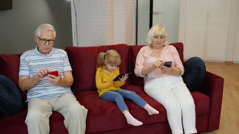 Grandparents-and-granddaughter-sit-on-the-couch-at-home-and-use-mobile-phones-and-digital-tablet