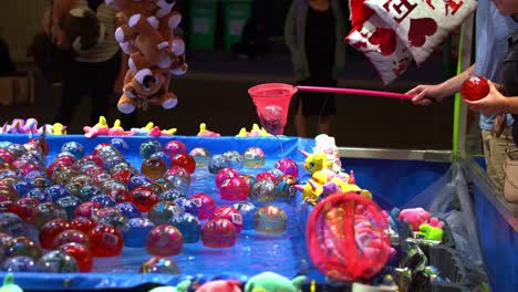 Popular-funfair-carnival-game,-man-using-a-net-and-catch-floating-ball-from-the-pool-with-winning-prize-for-his-girlfriend