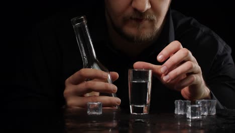 Alcoholic-man-pouring-up-frozen-vodka-from-bottle-into-shot-glass-with-ice-cubes-on-black-background