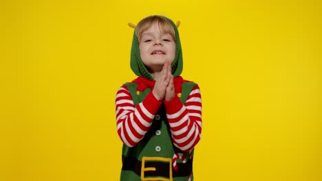 Kid-in-Christmas-elf-Santa-helper-costume-begs,-prays-about-gifts-on-New-Year.-Keeps-palms-together