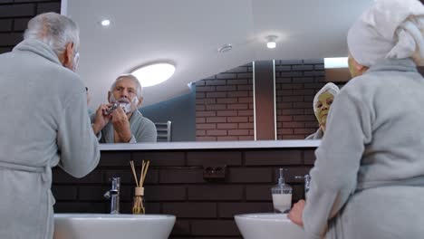 Senior-grandmother-applying-facial-mask-and-grandfather-shaving-with-manual-razor-blade-at-bathroom