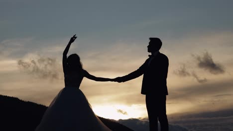 Newlyweds-silhouettes-bride,-groom-dancing-on-mountain-slope,-holding-hands,-wedding-couple-family