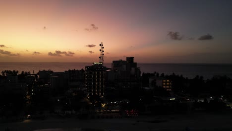 Cielo-Del-Atardecer-Brillando-Detrás-De-La-Isla-De-Maafushi-En-Vista-De-Silueta-De-Dron,-Estableciendo-Descendente