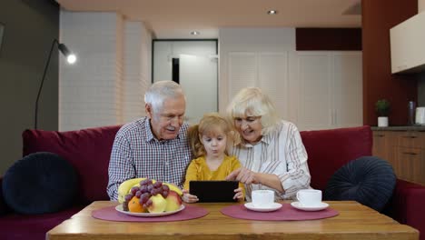 Senior-Caucasian-couple-with-cute-child-girl-granddaughter-using-tablet,-watching-funny-videos