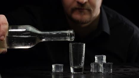 Bartender-pouring-up-frozen-vodka-from-bottle-into-shot-glass-with-ice-cubes-on-black-background