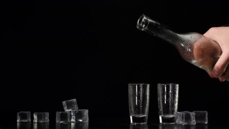 Hand-pours-vodka,-tequila-or-sake-from-bottle-into-shot-glasses-on-black-background-with-ice-cubes