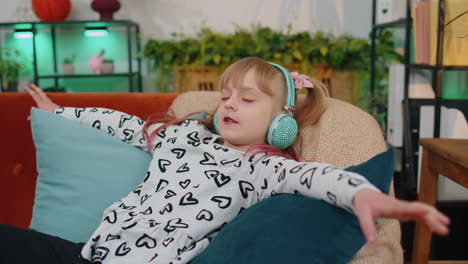 Little-children-girl-lying-on-sofa,-wearing-wireless-headphones,-listening-to-music,-singing-song