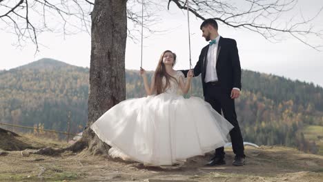 Newlyweds.-Caucasian-groom-with-bride-ride-a-rope-swing-on-a-mountain-slope