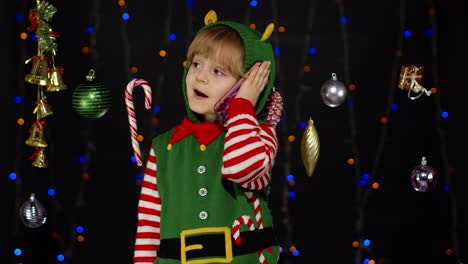 Kid-girl-in-Christmas-elf-Santa-Claus-helper-costume-making-congratulations-call-on-mobile-phone