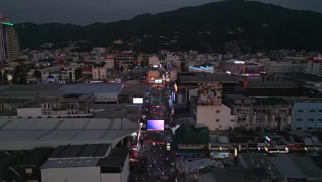 Forward-moving-aerial-shot-of-Bangla-Road-with-tourists-and-local-people-at-Patong-Beach-and-cityscape-at-background-during-sunset-in-Phuket,-Thailand