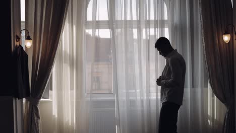 Man-in-jacket-fastens-buttons,-buttoning-white-shirt-preparing-to-go-out,-walking-near-window