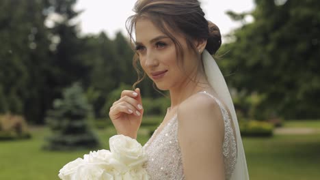 Beautiful-stylish-bride-in-white-wedding-dress-and-veil-holding-wedding-bouquet-in-hands-in-park