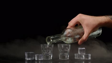 Hand-pours-vodka,-tequila-or-sake-from-bottle-into-shot-glasses-on-black-background-with-ice-cubes