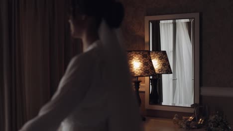 Lovely-bride-in-white-boudoir-dress-going-to-balcony-and-opens-curtains-in-hotel-room-or-apartment