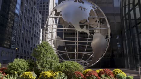 Steel-globe-sculpture-in-Chicago-with-a-pan-and-tilt-shot,-surrounded-by-urban-buildings-and-vibrant-flowers