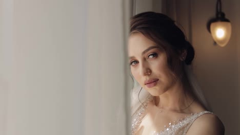 Bride-in-white-dress-staying-near-window-and-looking-at-camera,-wedding-morning-preparations-at-home