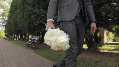 Groom,-young-man-businessman-in-green-summer-park-with-a-wedding-bouquet-in-hands,-wedding-day