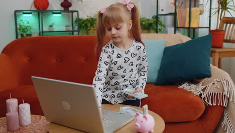 Little-kid-girl-counting-money-dollar-banknotes-for-future-needs-at-home-desk-with-laptop,-piggybank