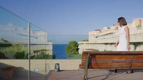 Female-brunette-in-long-white-dress-walk-to-glass-balcony-to-enjoy-view,-Spain